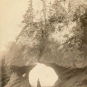 Photographie noir et blanc montrant une arche rocheuse sous laquelle se tient une femme en robe longue sur la plage
