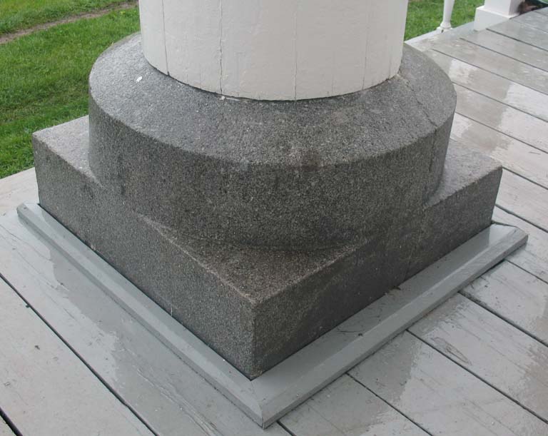 Photographie couleur d’une dalle de roche grise mouchetée, carrée à la base, cylindrique au sommet, supportant une colonne en bois blanche