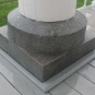 Photographie couleur d’une dalle de roche grise mouchetée, carrée à la base, cylindrique au sommet, supportant une colonne en bois blanche