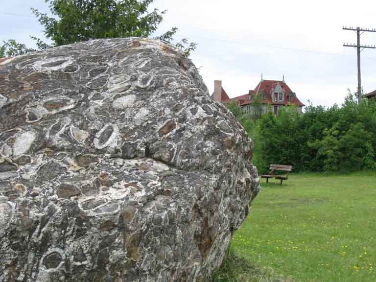 Photographie couleur d’un gros bloc de roche grise sur l’herbe verte avec un bâtiment à l’arrière-plan