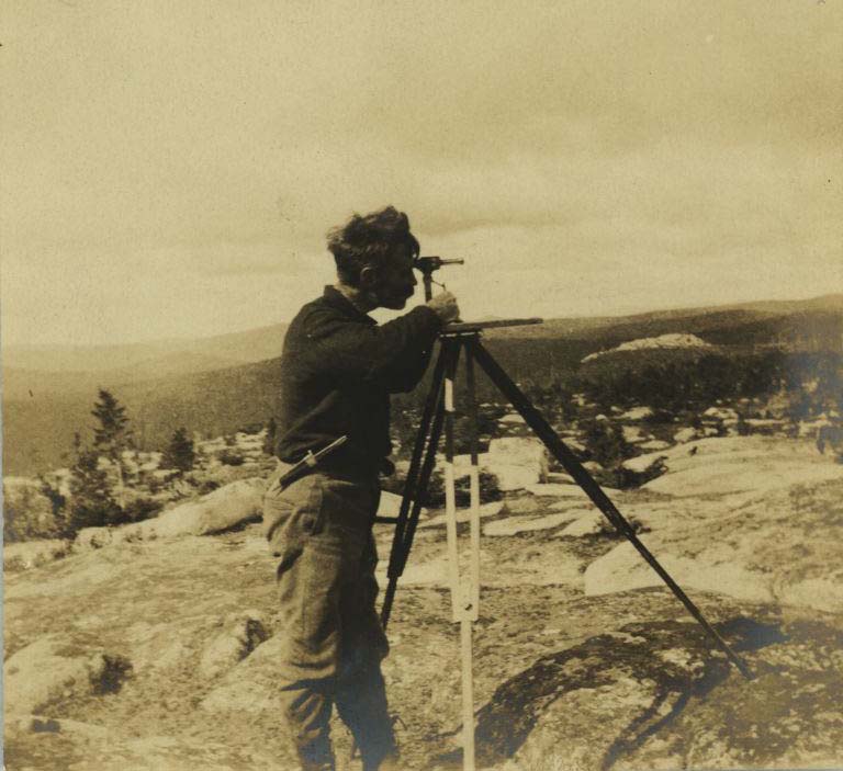 Black and white photograph of man standing on rocks with surveying equipment