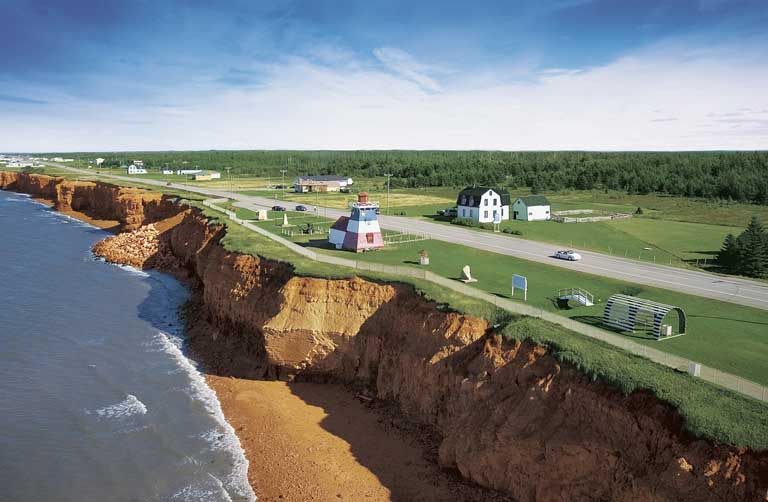 Colour photograph of a shoreline with brown cliffs and green fields on top with a lighthouse building